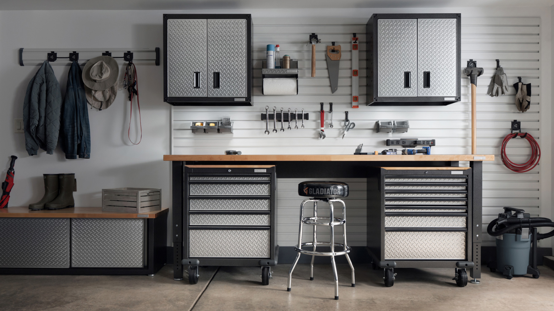 Un mur de garage avec deux armoires suspendues Gladiator. Autour, sur des crochets, se trouvent des outils et un organiseur pour vis, écrous et boulons, ainsi qu’un porte-rouleau d’essuie-tout Gladiator. Sous les armoires, on voit un établi Gladiator avec un tabouret. À côté, un aspirateur industriel et un organiseur à chaussures Gladiator sur lequel sont posées des bottes. Des vestes et des chapeaux sont suspendus à un système Gladiator GearTrack