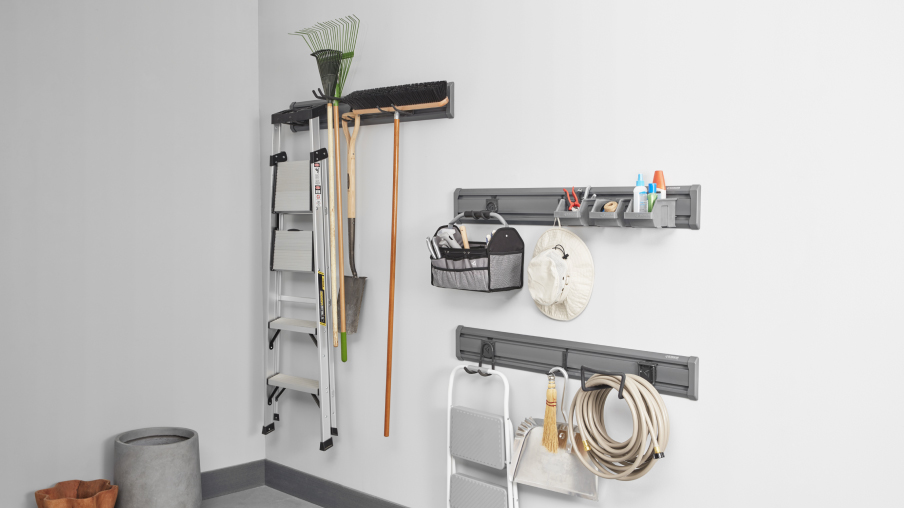 Sur des solutions de rangement Gladiator comme des systèmes GearTrack sont suspendus une échelle, un râteau, un balai, un chapeau, un boyau d’arrosage et un petit escabeau.