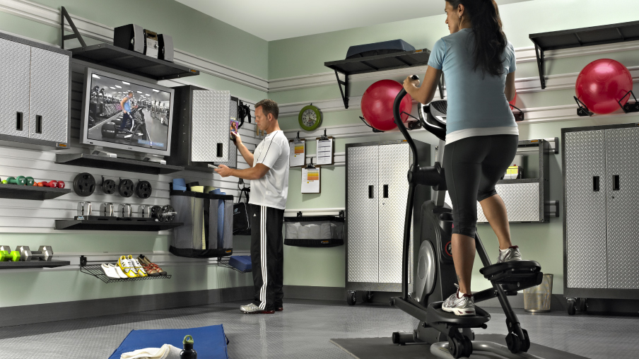 A home gym where a man looks inside a Gladiator Cabinet, and a woman works out on an elliptical. Also in the home gym are two Gladiator Storage Units, Gladiator Cabinets, and dumbbells and weights are on shelves.