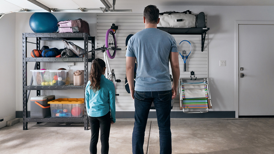Un homme et une jeune fille se tiennent devant un mur bien organisé de leur garage. On y voit une étagère Gladiator remplie d’articles, du rangement en hauteur et des objets comme un vélo et une raquette de tennis accrochés au mur