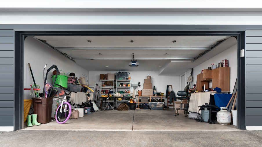Un garage avec la porte ouverte. L’intérieur est rempli d’articles rangés le long du mur. À l’arrière se trouvent deux étagères, et parmi les objets, on aperçoit un vélo, une bonbonne de propane, une chaise et plus encore