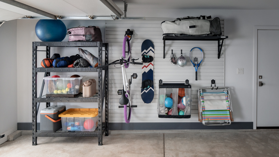 Le long d’un mur de garage se trouvent une étagère Gladiator, du rangement en hauteur Gladiator et des articles comme un vélo, une planche à neige et une raquette de tennis accrochés au mur. On y voit aussi un panier Gladiator rempli de ballons