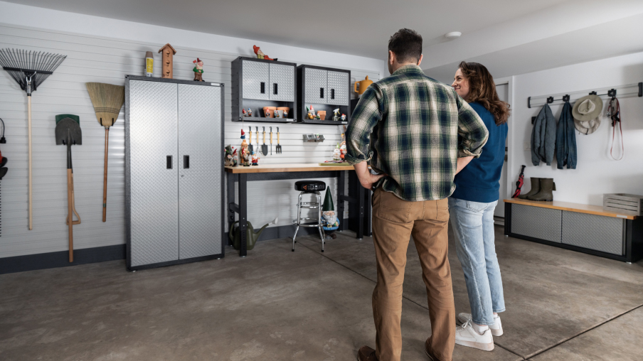 Un homme et une femme se tiennent devant leur garage bien organisé. On y voit une unité de rangement Gladiator, deux armoires Gladiator et un établi Gladiator. Sur un mur, des chapeaux et des manteaux sont accrochés à un Gladiator GearTrack, et sur un autre, un râteau, une pelle et un balai sont suspendus