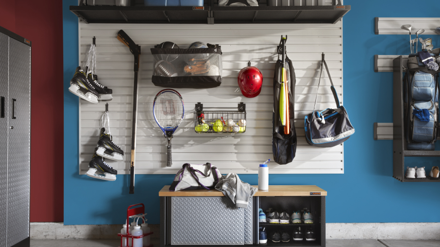Un mur de garage organisé avec des solutions de rangement Gladiator, comme des bacs et des crochets. Parmi les articles suspendus, on trouve des patins, des raquettes de tennis, des ballons, un casque et un bâton de hockey.