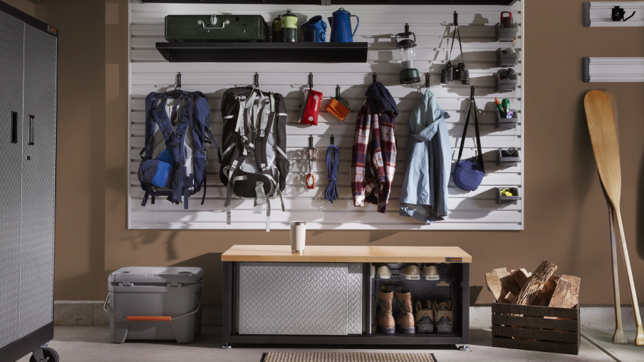 Dans un garage se trouve une unité de rangement Gladiator à côté d’un mur où des articles sont organisés sur des crochets et des tablettes Gladiator. On y voit des sacs à dos, des vestes et des bacs remplis d’articles. Sous le mur se trouve un organiseur à chaussures Gladiator. Deux rames sont appuyées contre le mur.