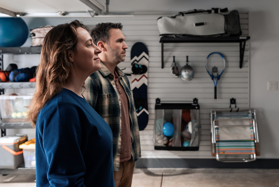 Un homme et une femme regardent un mur. Derrière eux se trouvent des articles comme une planche à neige, une raquette de tennis et un casque sont accrochés au mur. On voit aussi un panier Gladiator rempli de ballons et des étagères Gladiator où sont rangés de nombreux articles