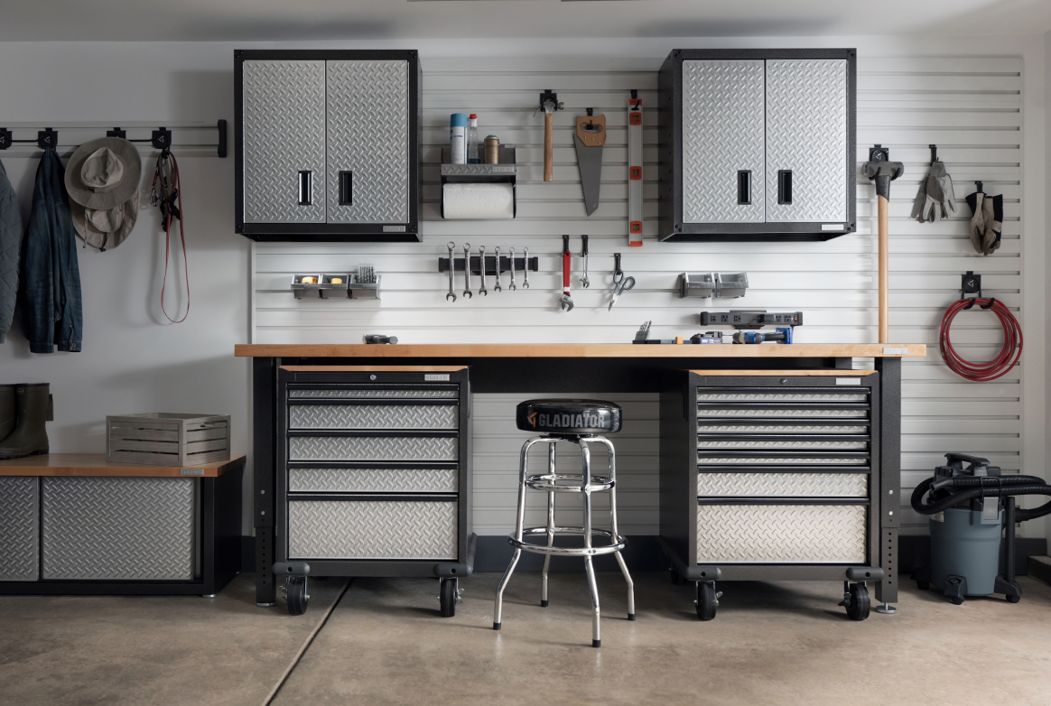 Le mur d’un garage organisé avec des systèmes de rangement Gladiator. On y voit deux armoires Gladiator, un établi Gladiator et plus. Des outils, dont une scie et un niveau, sont accrochés au mur à l’aide de crochets Gladiator. À côté, on voit une veste et des chapeaux suspendus à un système Gladiator GearTrack