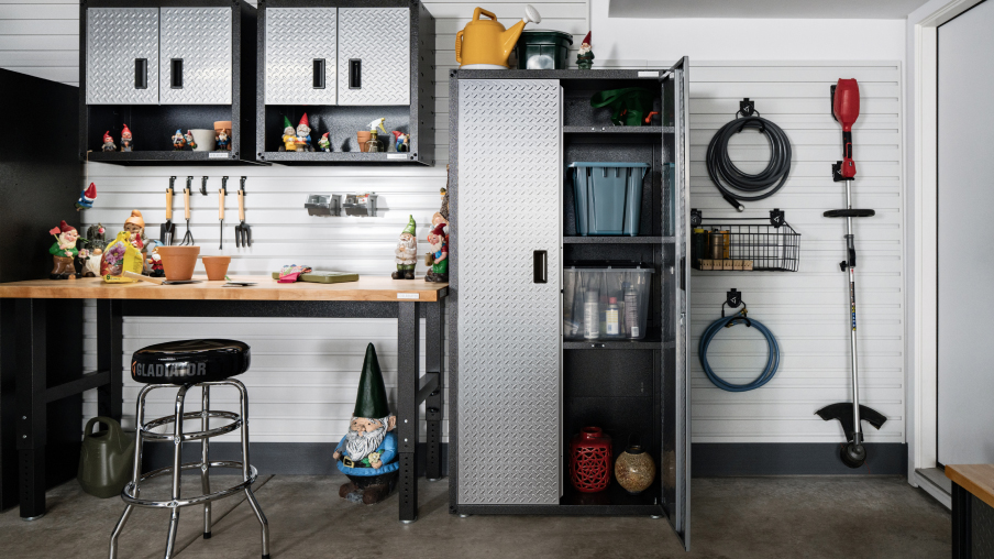 Un garage avec une unité de rangement Gladiator dont la porte est ouverte. À côté se trouve un établi Gladiator et au-dessus de l’établi, deux armoires Gladiator. Sur le mur, deux boyaux d’arrosage et un coupe-herbe sont accrochés à l’aide de crochets Gladiator.