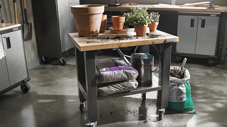  milieu d'un garage se trouve un établi Gladiator sur lequel sont posés des pots de fleurs. Sur l'étagère du bas sont posés des sacs de terreau et un arrosoir. Derrière, on voit deux armoires Gladiator