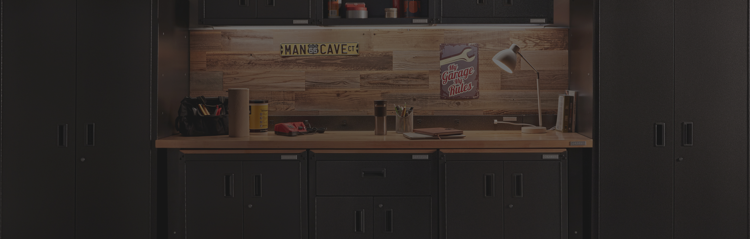 A Gladiator Workbench in a garage. On the wall is a sign that reads, "MAN CAVE" and a poster of a wrench that reads, My Garage My Rules". On the desk is a lamp turned on, a roll of paper towel, a yellow container, a toolbelt with various tools, notebooks and books