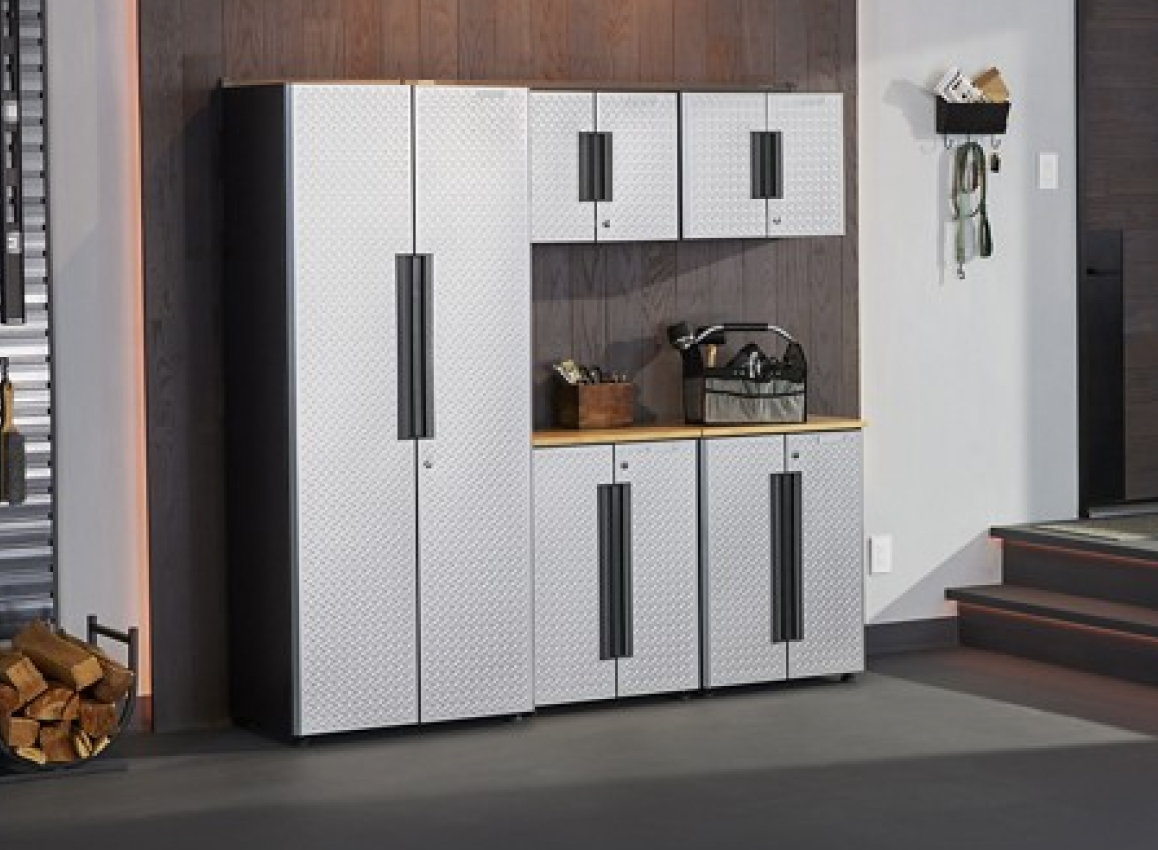 A Garage with Gladiator Storage Units. On top of the cabinets are a toolbox holding items like a paintbrush and a brown box. On the floor are logs in a holder. On the wall are items in a Gladiator Bin and keys and an extension cord hang from Gladiator Hooks