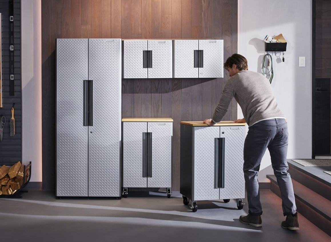 A man sets up a Gladiator Flex Cabinet System. He's rolling a cabinet next to another cabinet in a garage. On the wall are a bin and hooks with various items. On the floor is a basket with logs. On the wall behind the logs are hooks with levers and a brush