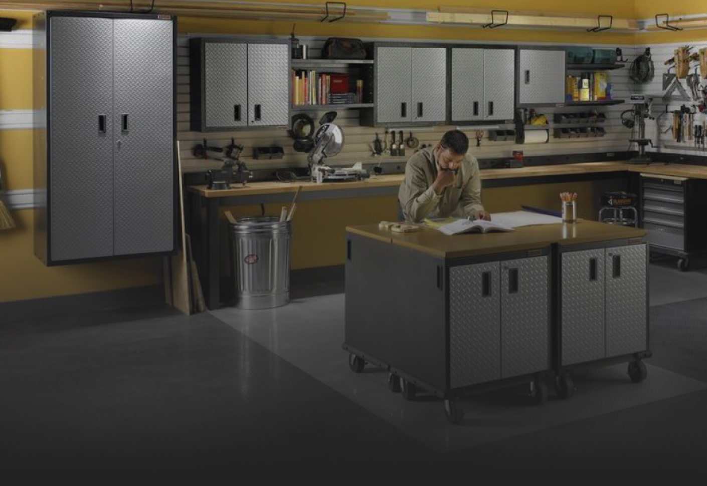 A man reads a manual at a Gladiator Workbench next to a full pencil holder. Gladiator Cabinets are behind him on the wall and there's a Gladiator Mobile Tool Storage Chest. Tools and automatic tools are along the workbench in the background and various items hang on Gladiator Hooks and in Gladiator Bins