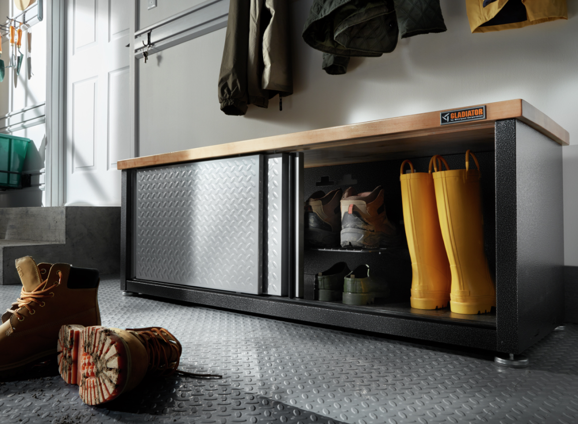 Un banc de rangement pour chaussures Gladiator. La porte est ouverte et révèle des chaussures et des bottes de pluie jaunes. On voit des vestes suspendues au-dessus et une paire de bottes au devant