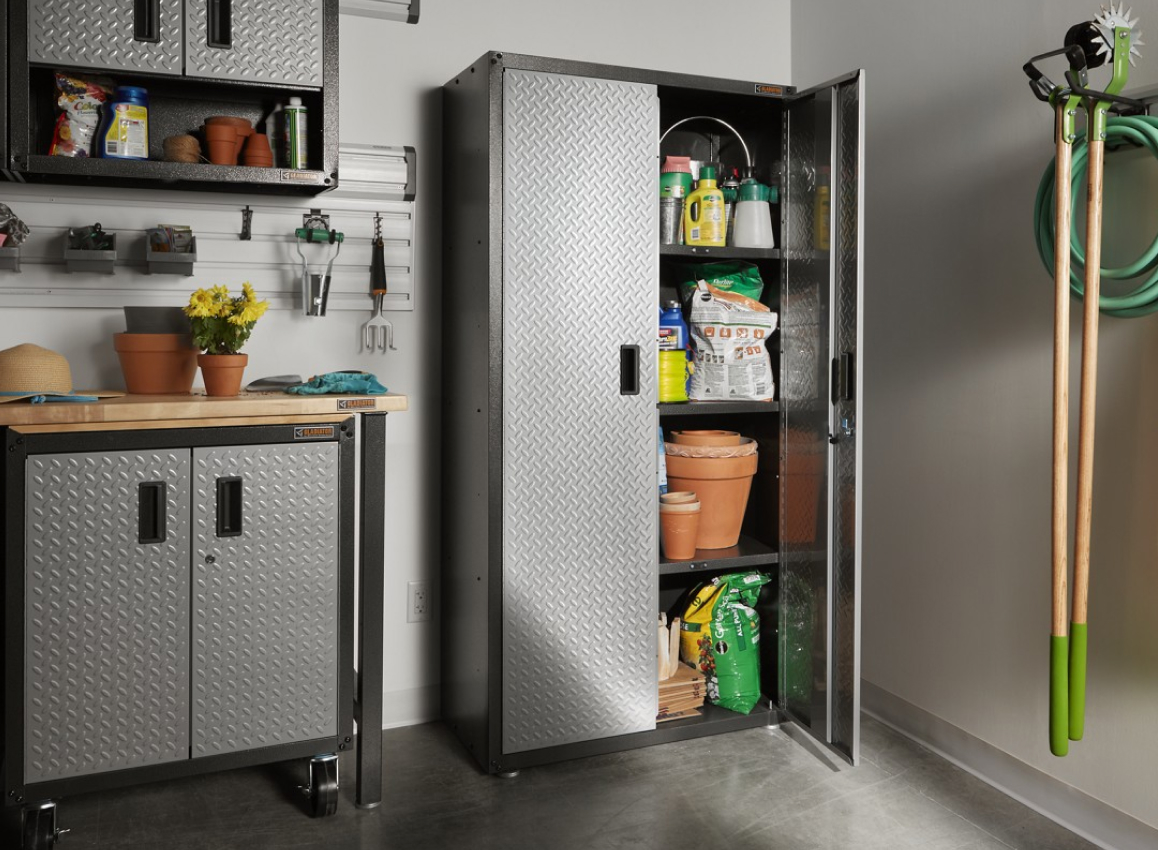 Une armoire à outils Gladiator avec une porte est ouverte. À l'intérieur se trouvent un sac de terre, des articles de jardinage, des pots pour les plantes et d'autres objets. À côté, une boîte à outils Gladiator avec des plantes en pot sur le dessus et des outils de jardinage sont accrochés au mur