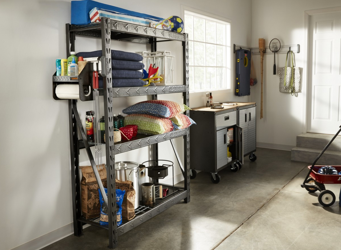 A garage with a Gladiator Shelving Unit with pillows, watering cans, a Gladiator Clean-up Caddy and other items. There's also a red wagon, a Gladiator Workbench, and items on a Gladiator GearTrack.