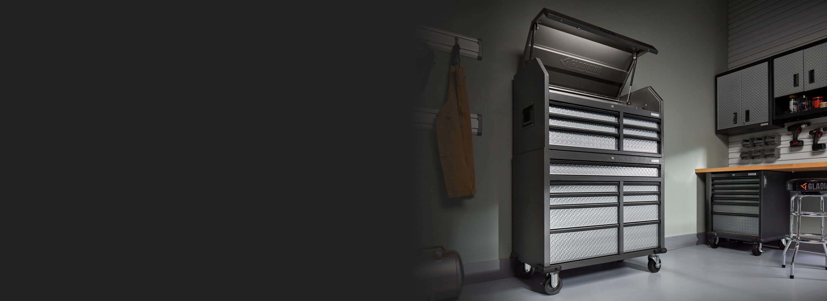 A Gladiator Tool Storage Chest Combo with the top opened. Nearby, work attire hangs on a Gladiator GearTrack System. On the other side of the chest are Gladiator Modular Units, a stool and tools like drills hanging on Gladiator Hooks on the wall