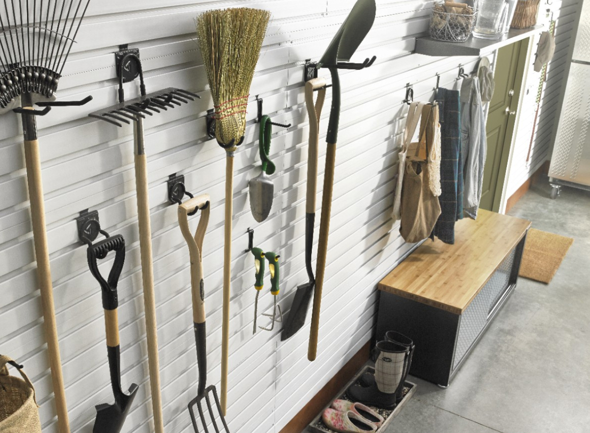 Hanging on a Gladiator GearTrack system are gardening tools like a rake and shovels, among other items. Nearby, aprons hang above a Gladiator Storage Shoe Bench and below a shelf of items
