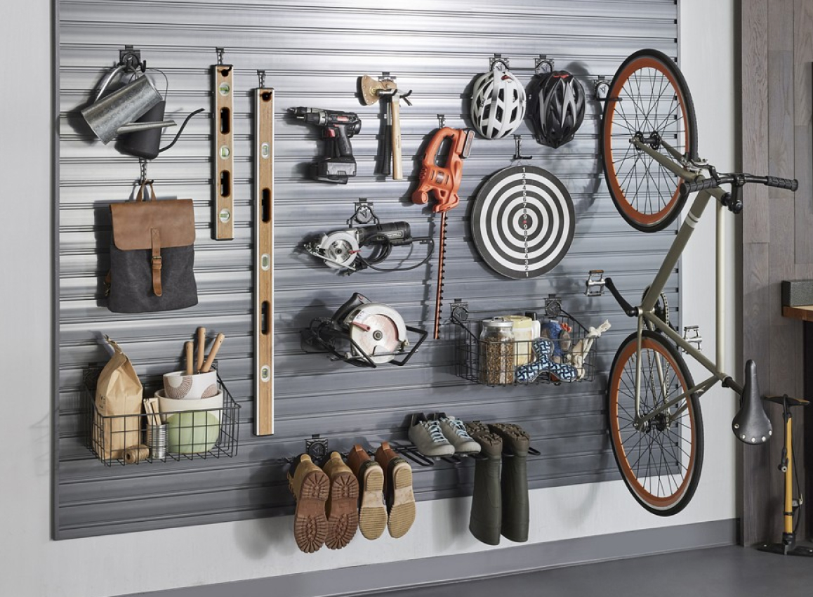 Hanging on a Gladiator GearTrack system are a watering can, a bag, a basket with items, two levels, tools like drills, shoes, boots, a bullseye, bike helmets, a bike and another basket filled with items
