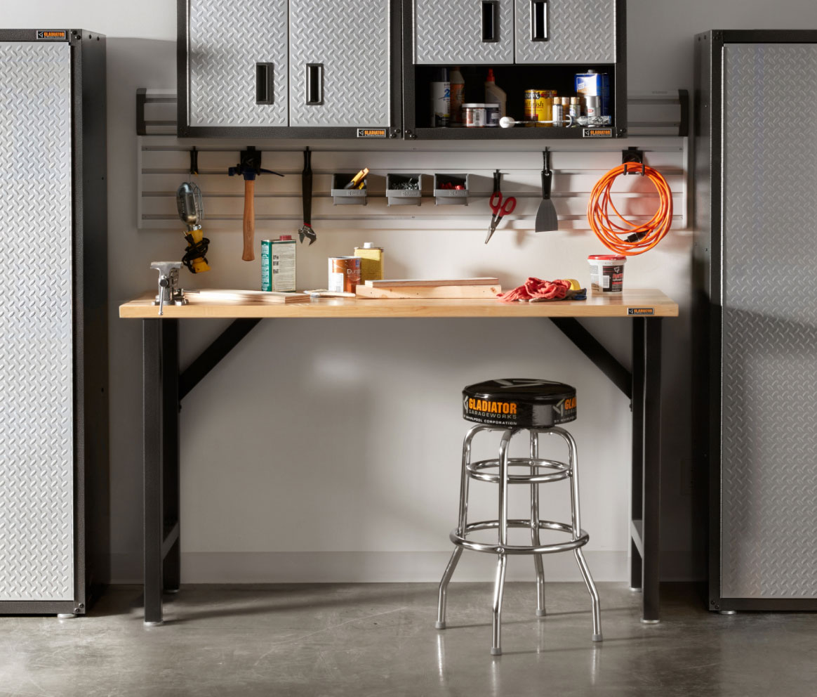 A garage with a Gladiator Workbench and a stool. On the workbench are various items and on the wall are Gladiator Bins and Gladiator Hooks where scissors, a hammer, a wrench and an extension cord hang. Next to the workbench are two Gladiator Full-Size Cabinets