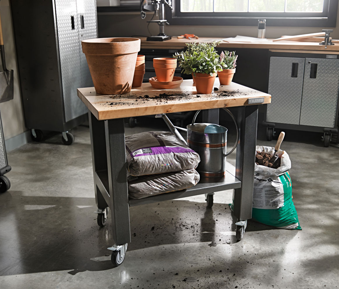 A Gladiator Mobile Workstation with potted plants on top. On the below shelf are bags of soil and a watering can. Next to it is an opened bag of soil with a gardening tool sticking out