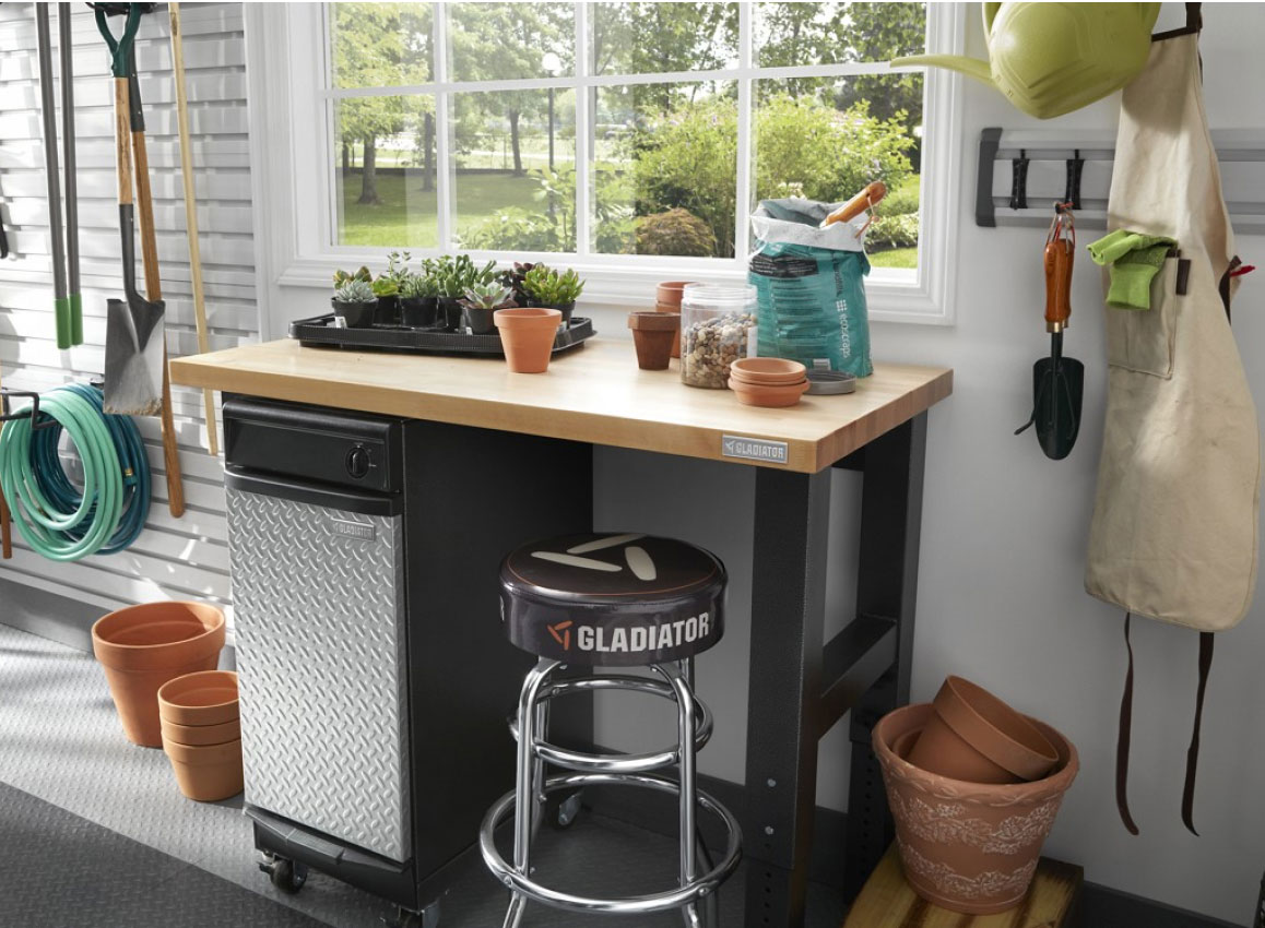 A garage with a Gladiator Workbench with a Gladiator Modular Cabinet underneath. There's a stool and several plant pots nearby. On the wall on a Gladiator GearTrack is a gardening shovel and apron. On the far wall hangs a garden hose and gardening tools