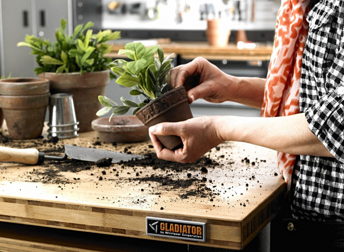 Someone in a black and white checkered shirt touches a potted plant in front of a Gladiator Workbench that's covered in soil. On the workbench are a stack of pots and a potted plant and a garden tool