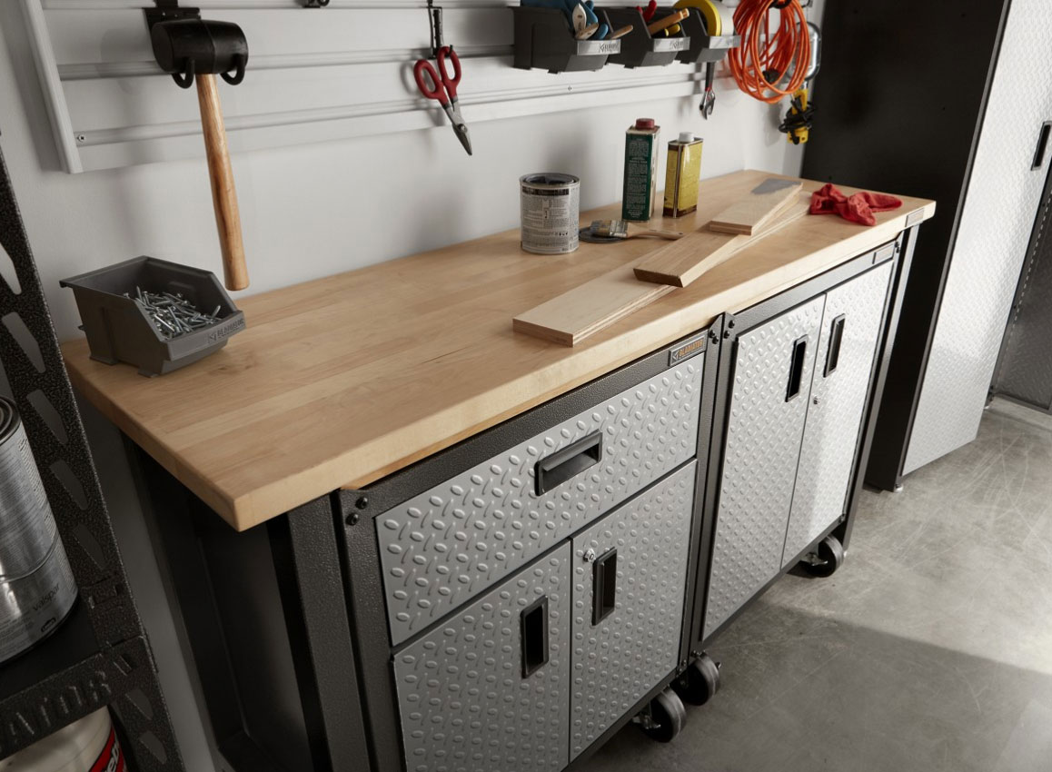 A Gladiator Workbench and Gladiator Full-Size Cabinet. On the workbench are wood planks, a Gladiator bin with nails, a rag and cans. Hanging on the wall are a mallet, scissors, wrench and extension cord