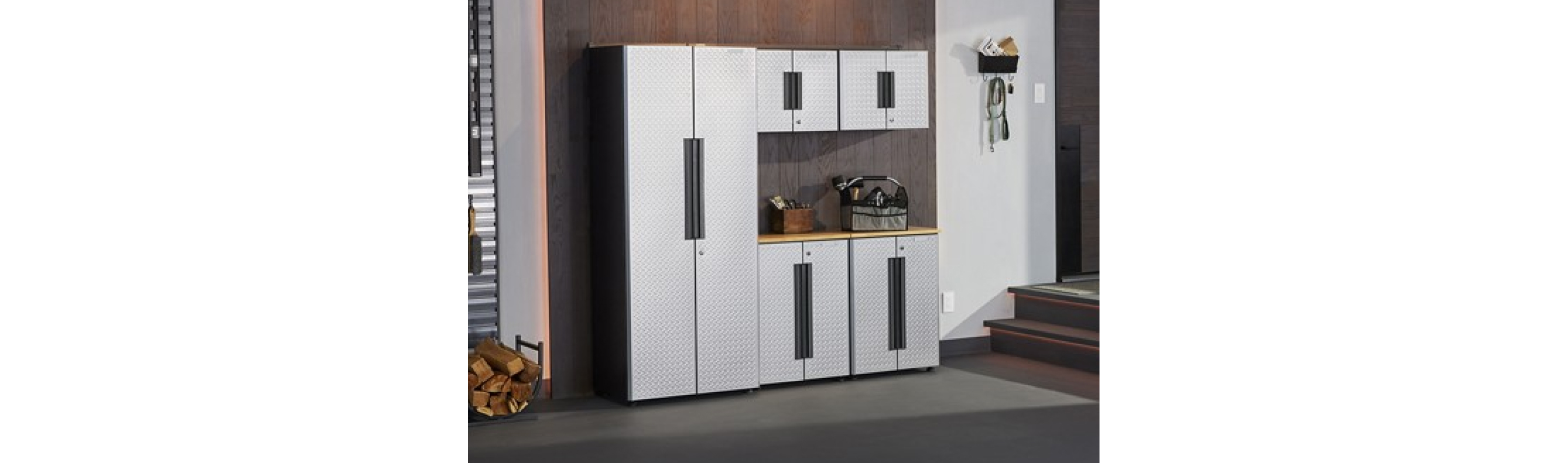 A Garage with Gladiator Storage Units. On top of the cabinets are a toolbox holding items like a paintbrush and a brown box. On the floor are logs in a holder. On the wall are items in a Gladiator Bin and keys and an extension cord hang from Gladiator Hooks