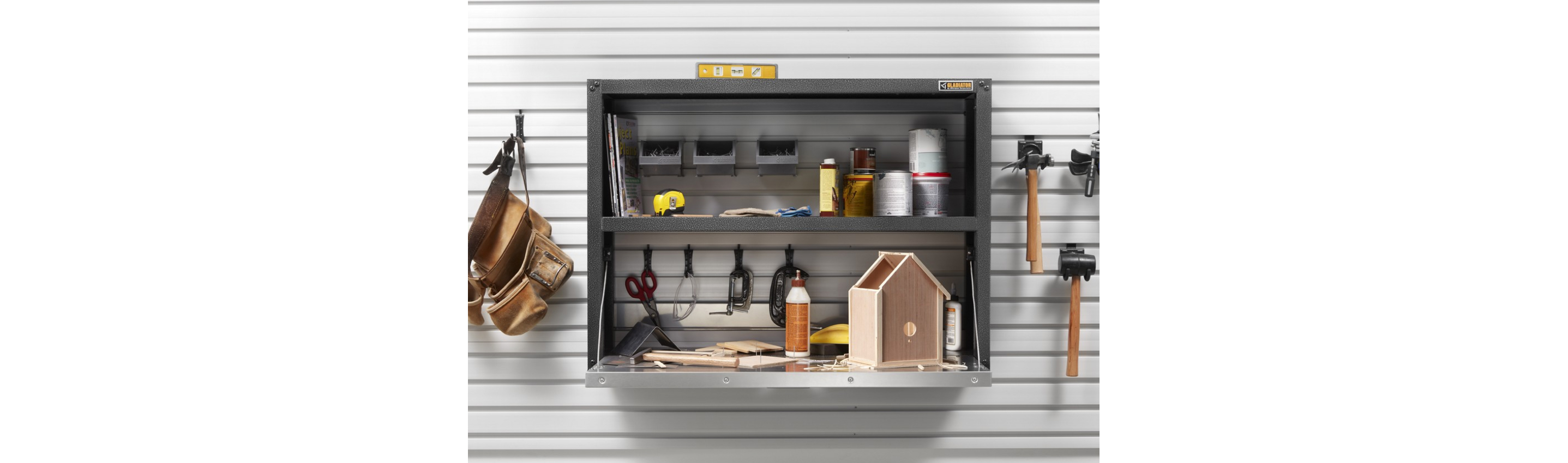 On a wall, hanging on Gladiator Hooks are a toolbelt, mallet, hammer and another item. In a Gladiator Shelving Unit are an almost completed birdhouse, glue, scissors, bins, cans, tape measure and more