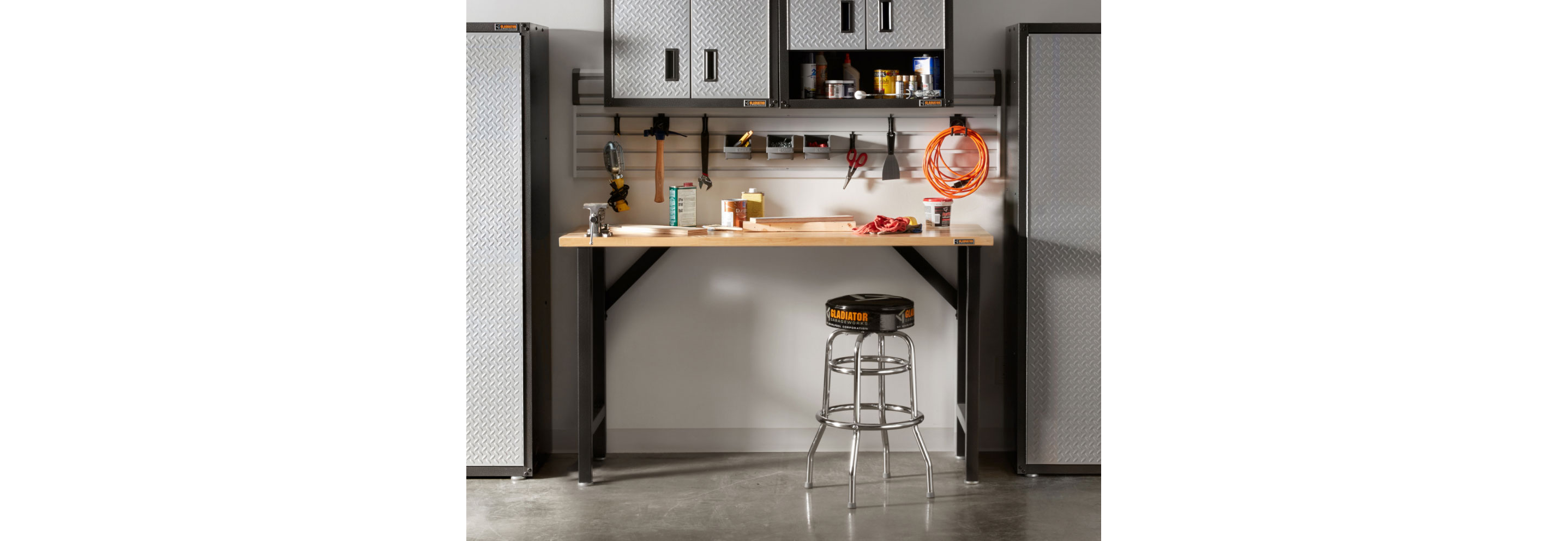 A garage with a Gladiator Workbench and a stool. On the workbench are various items and on the wall are Gladiator Bins and Gladiator Hooks where scissors, a hammer, a wrench and an extension cord hang. Next to the workbench are two Gladiator Full-Size Cabinets