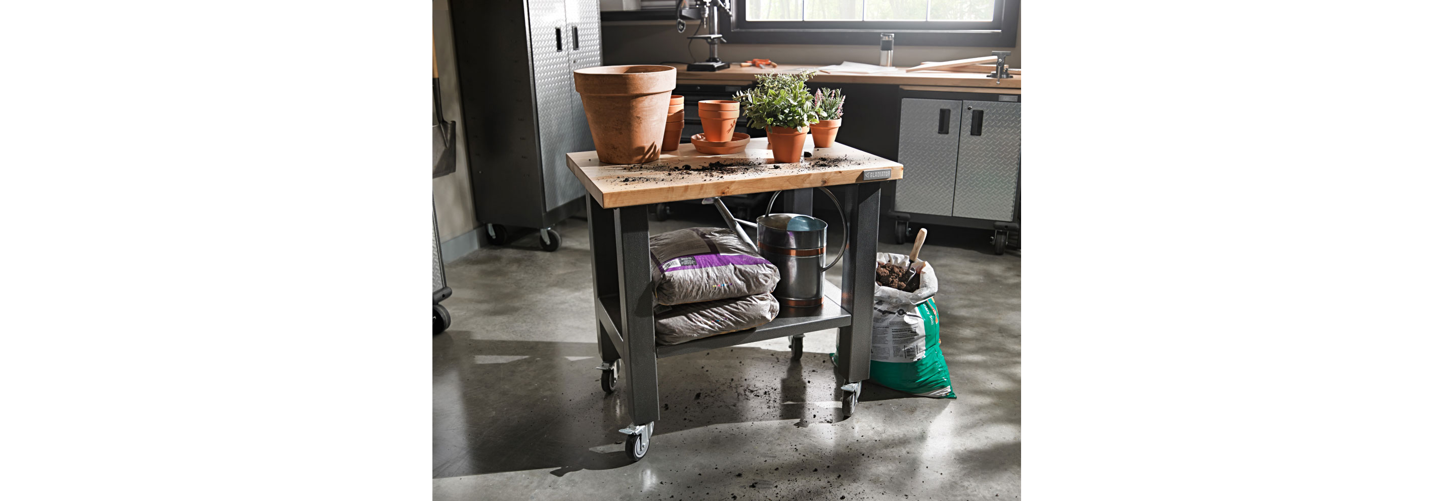 A Gladiator Mobile Workstation with potted plants on top. On the below shelf are bags of soil and a watering can. Next to it is an opened bag of soil with a gardening tool sticking out