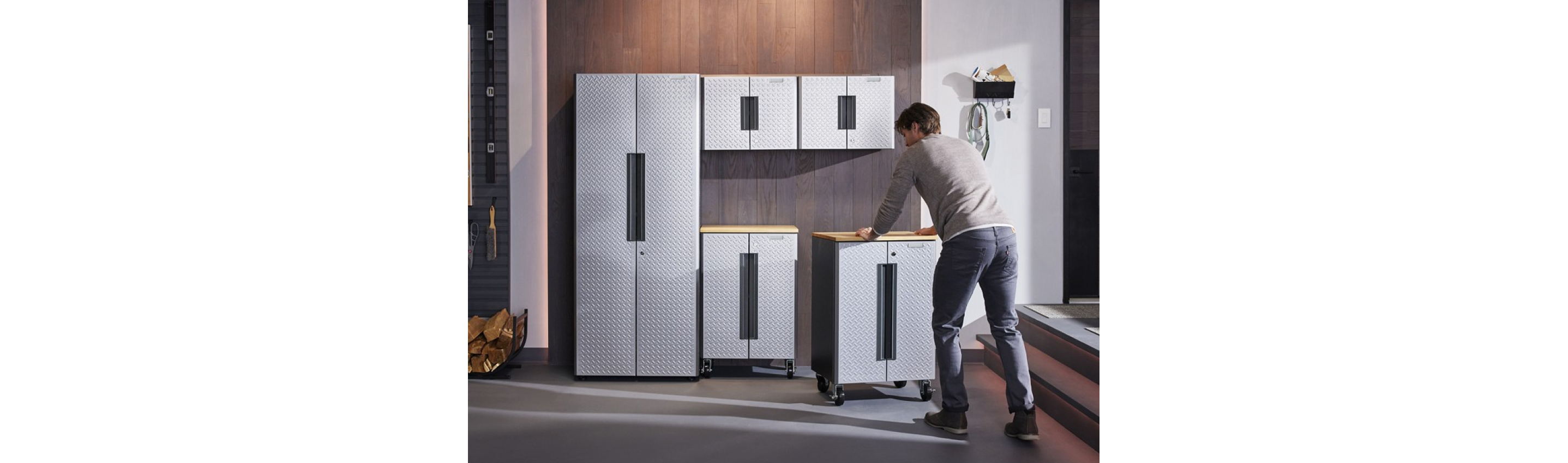  A man sets up a Gladiator Flex Cabinet System. He's rolling a cabinet next to another cabinet in a garage. On the wall are a bin and hooks with various items. On the floor is a basket with logs. On the wall behind the logs are hooks with levers and a brush
