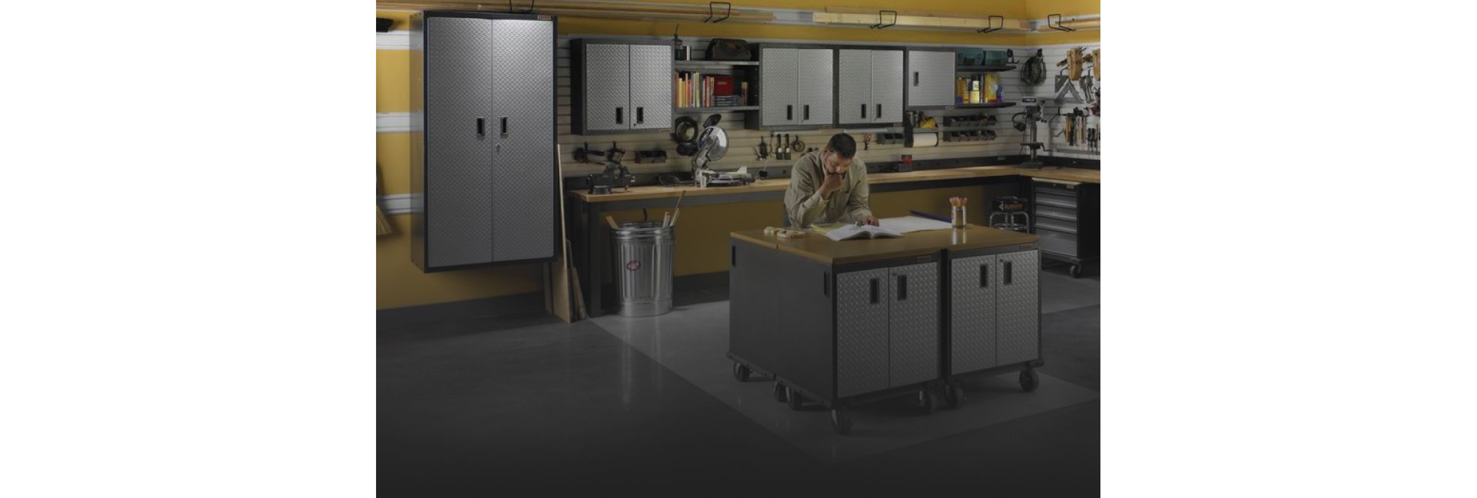 A man reads a manual at a Gladiator Workbench next to a full pencil holder. Gladiator Cabinets are behind him on the wall and there's a Gladiator Mobile Tool Storage Chest. Tools and automatic tools are along the workbench in the background and various items hang on Gladiator Hooks and in Gladiator Bins