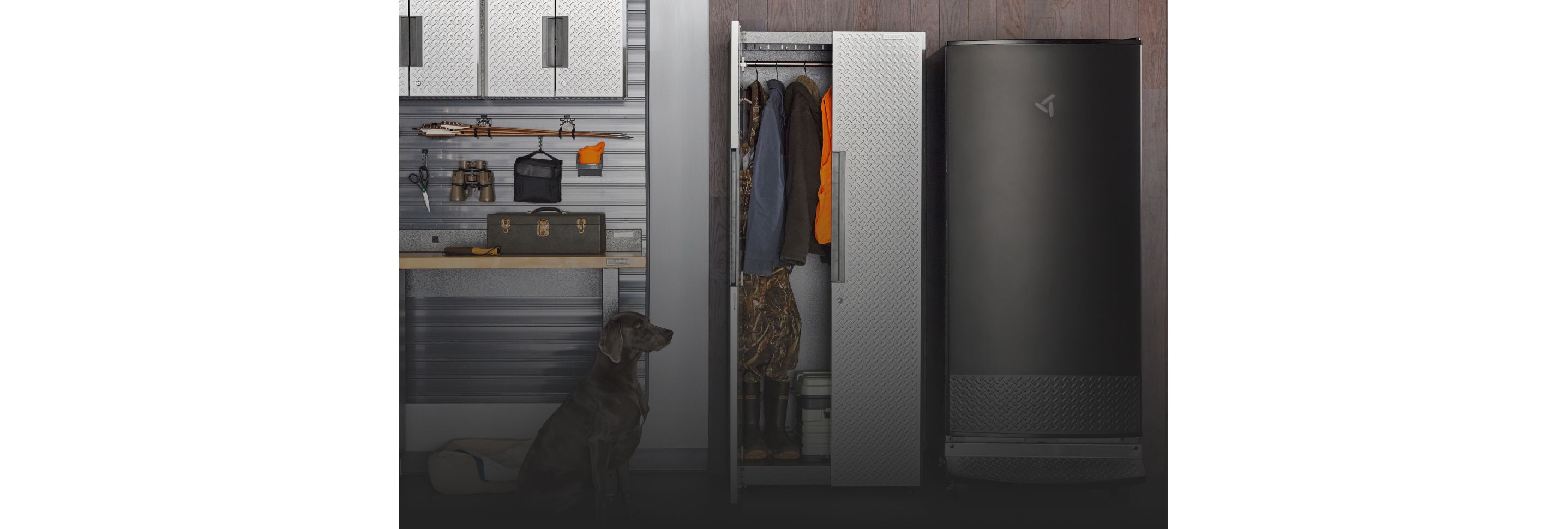 A garage with a sitting dog. A Gladiator Cabinet with one door opened. Inside are hanging jackets. Next to the cabinet is a Gladiator Garage Refrigerator