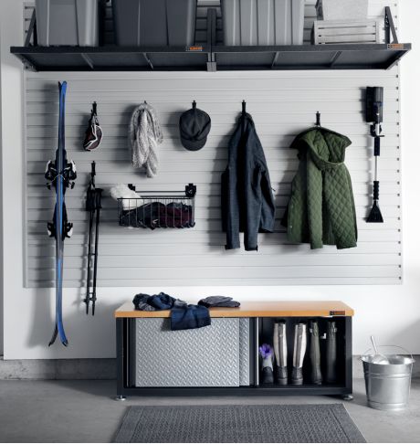 On a wall, hanging on Gladiator Hooks and in Baskets are skis, ski goggles, ski poles, jackets, winter items, a hat and an ice scraper. Below the hanging items is a Gladiator Storage Unit with an open sliding door revealing pairs of boots