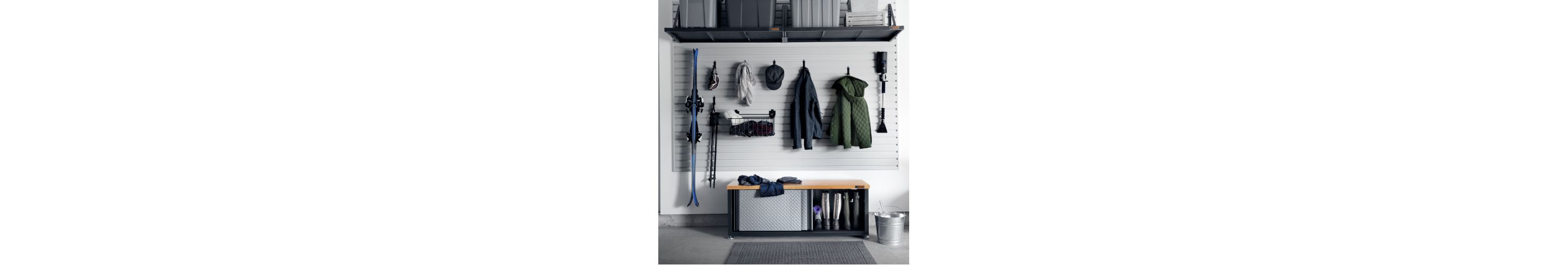 On a wall, hanging on Gladiator Hooks and in Baskets are skis, ski goggles, ski poles, jackets, winter items, a hat and an ice scraper. Below the hanging items is a Gladiator Storage Unit with an open sliding door revealing pairs of boots
